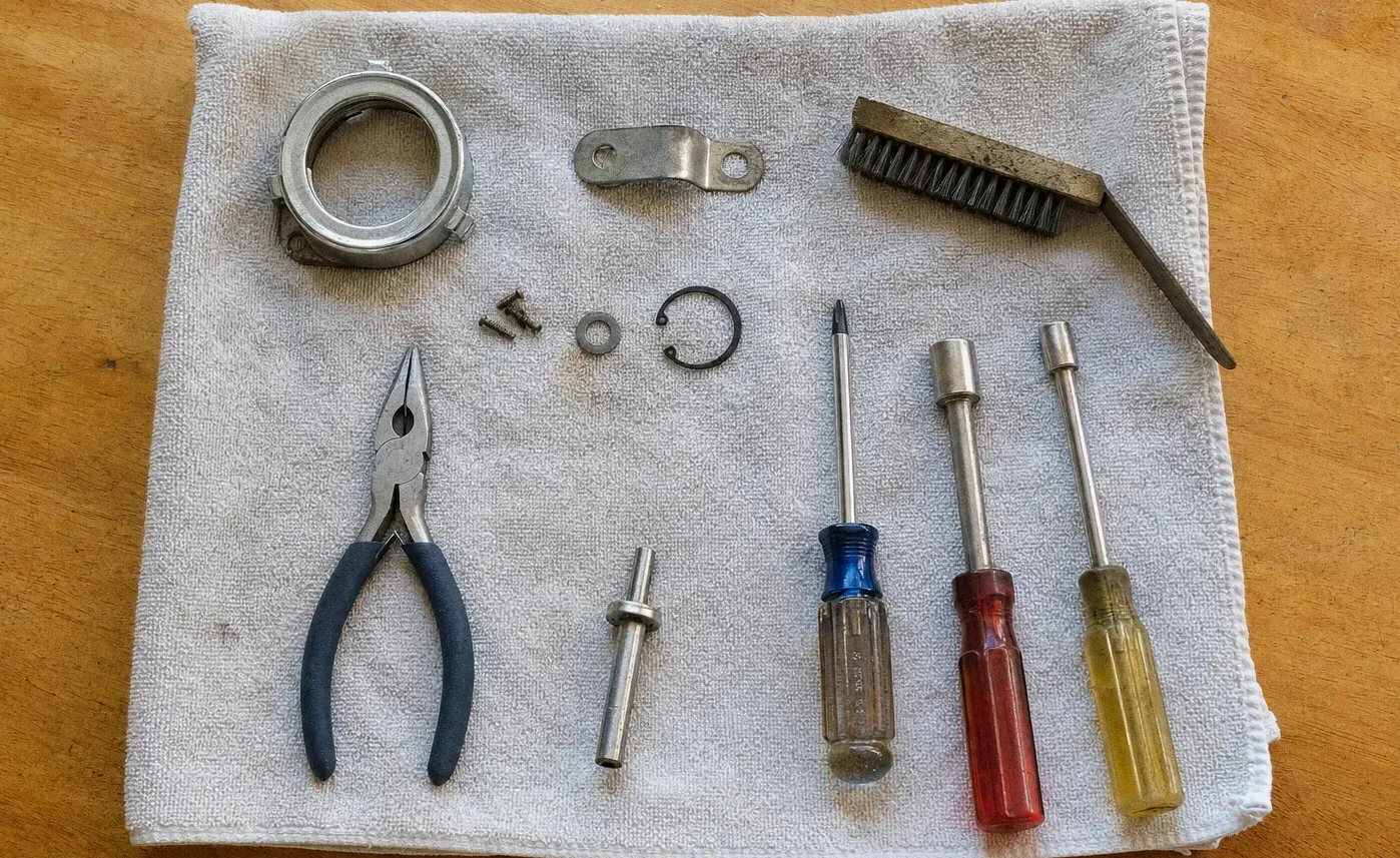 A burner ring, bracket, retaining clip and screws laid out on a towel beside a wire brush, pliers, a nut driver and three screwdrivers
