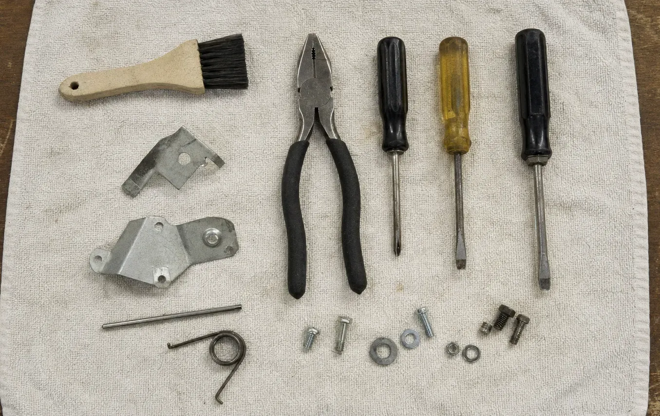 A wooden-handled brush, pliers and three screwdrivers laid out on a towel beside two hinge plates, a spring, screws and washers